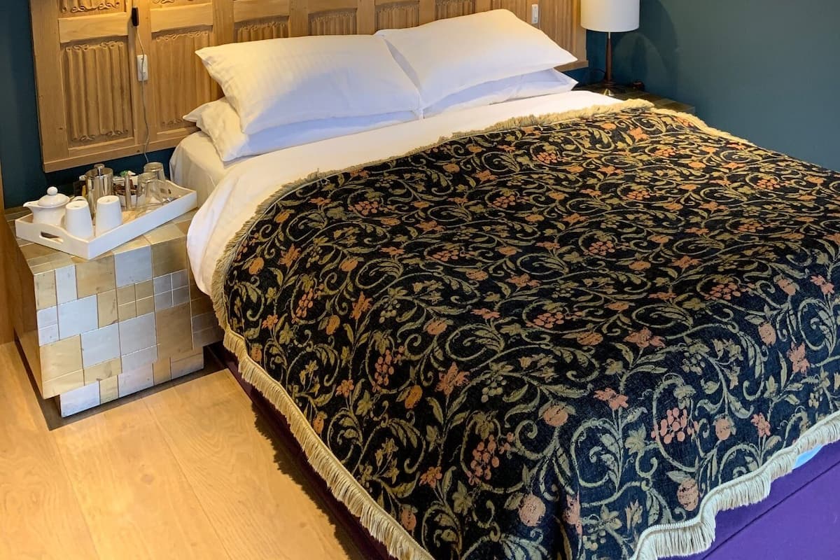 A neatly made bed with a patterned bedspread, a bedside table with a tray of toiletries, and a wall-mounted lamp.