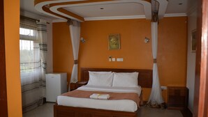 A bedroom with a wooden bed, white bedding, a bedside table, a nightstand, a wall-mounted light, and a small white appliance. - Le Savanna Country Lodge And Hotel (Kisumu)