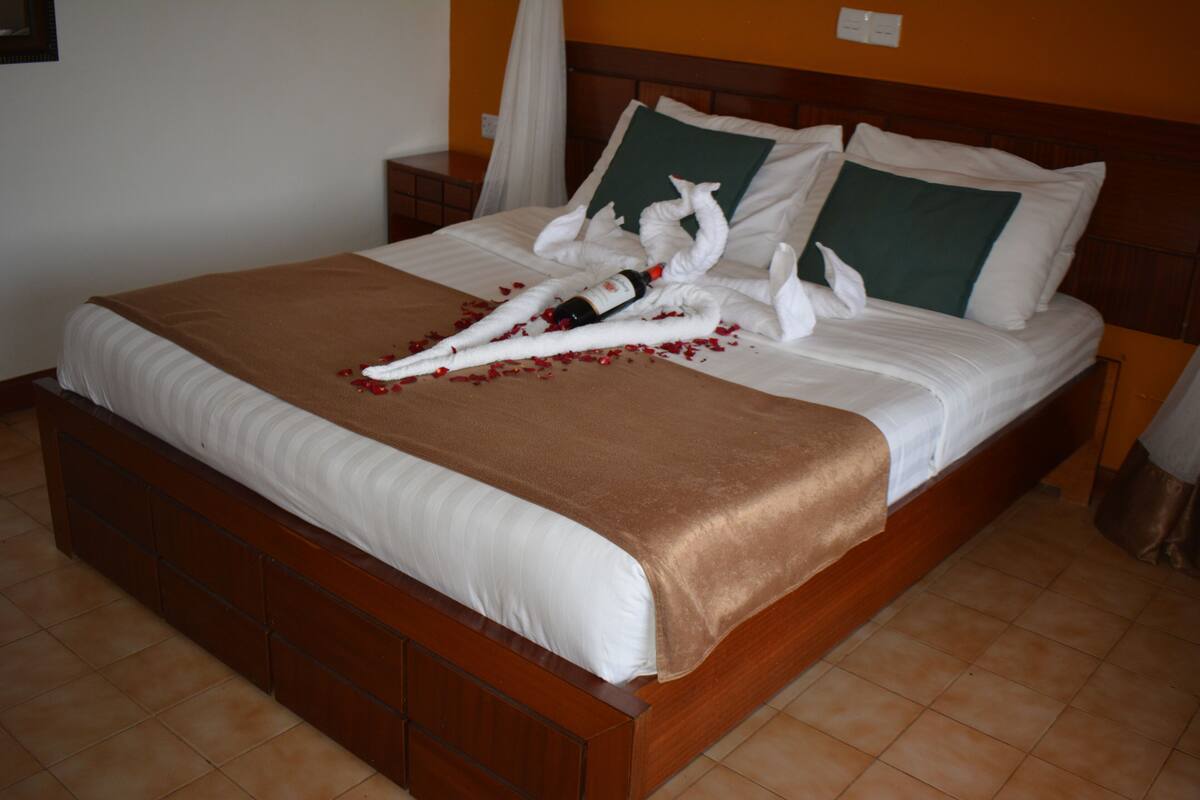 Una cama bien hecha con una manta marrón, almohadas blancas y un arreglo decorativo de toallas blancas y pétalos de rosa.