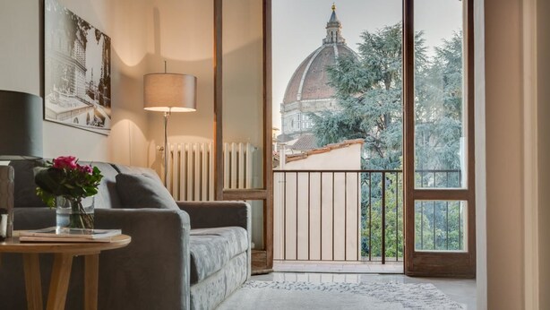 A living room with a sofa, a small table with a vase, a lamp, and a view of a dome through a balcony. - Alfani Views (Florence)