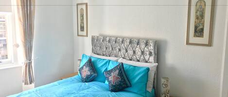 Cama bem arrumada com cabeceira estampada, colcha azul e almofadas decorativas. Tem uma mesinha de cabeceira com um vaso e um quadro na parede.