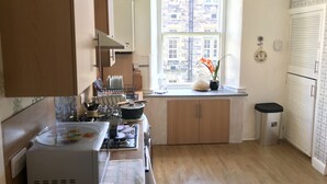 A compact kitchen with a window, wooden cabinets, a stove, and a trash bin. - 233 Leith Apartment (Edinburgh)