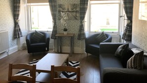 A living room with two sofas, a coffee table, and a chair. There are two windows with curtains, a radiator, and a small table with a potted plant. - 233 Leith Apartment (Edinburgh)