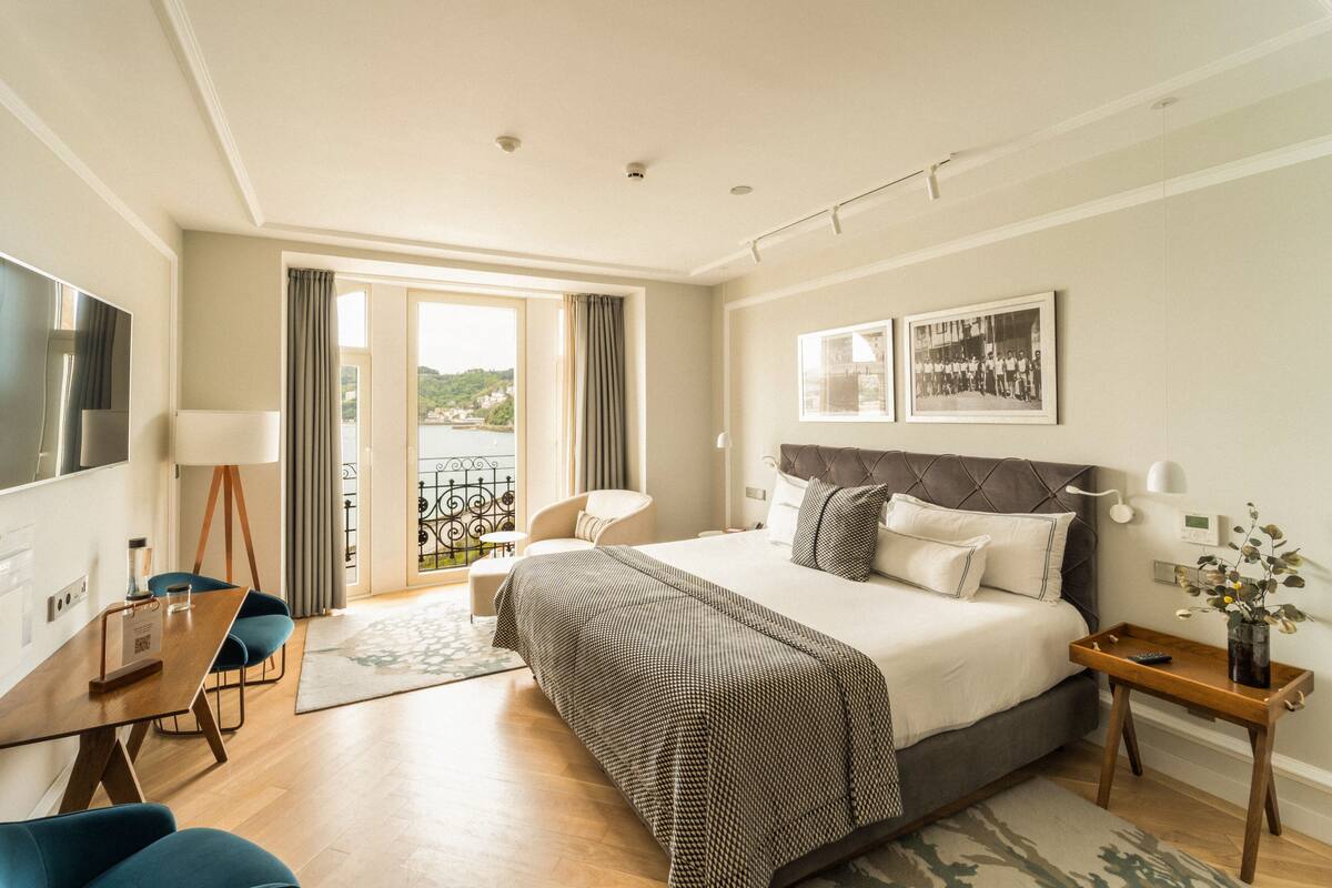 A hotel room with a large bed, a desk with a chair, a TV, and a balcony with a view of the sea.