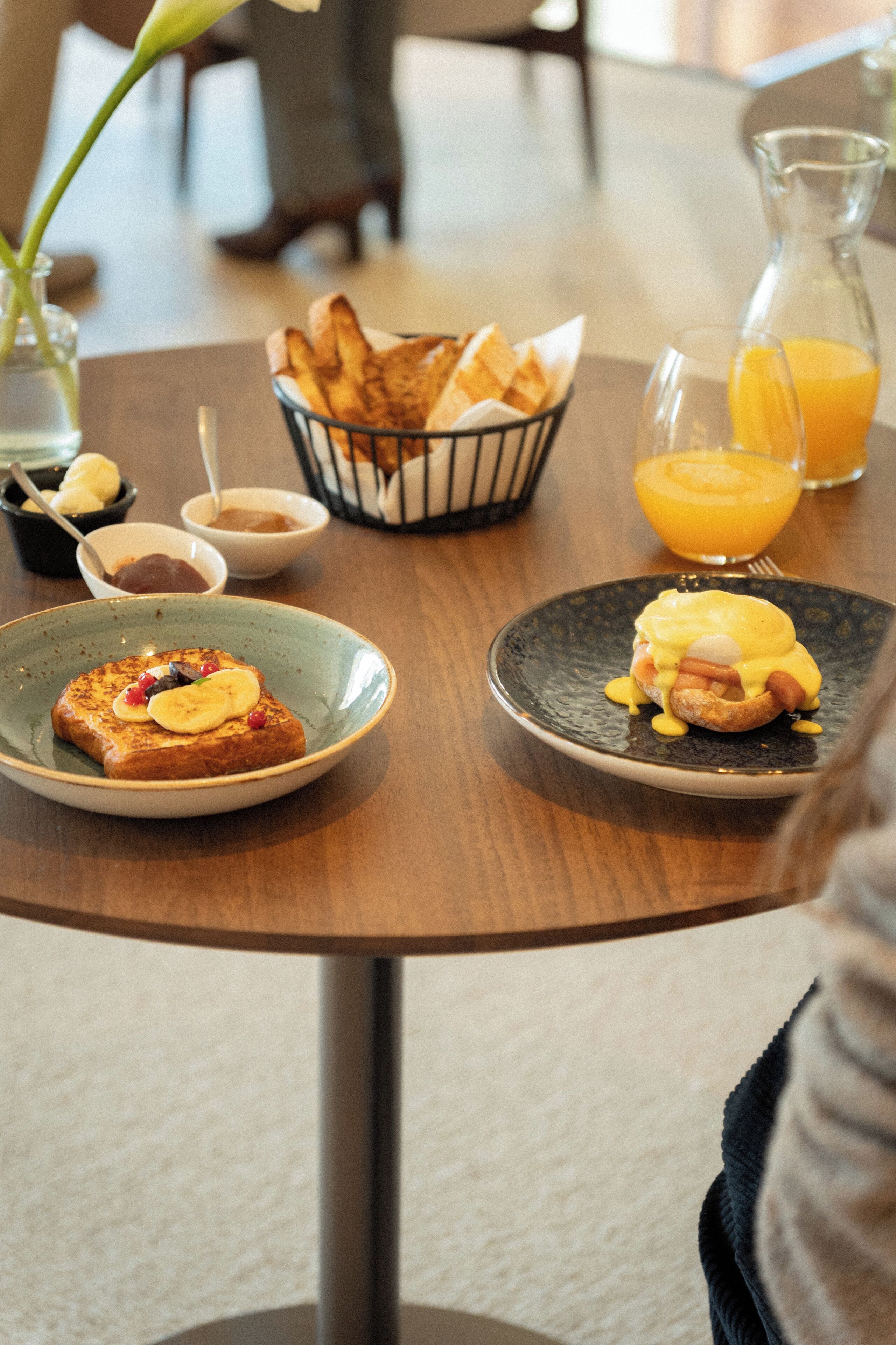 Desayuno con tostadas, huevos y fruta en platos, una cesta de pan y una jarra de jugo de naranja sobre una mesa de madera.