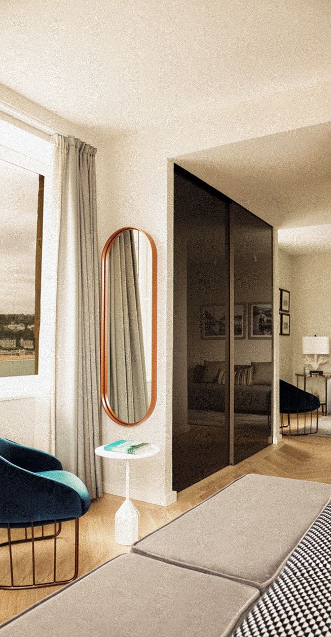 Una habitación de hotel moderna con un gran ventanal corredero, un sillón azul, una mesita redonda y un espejo grande.