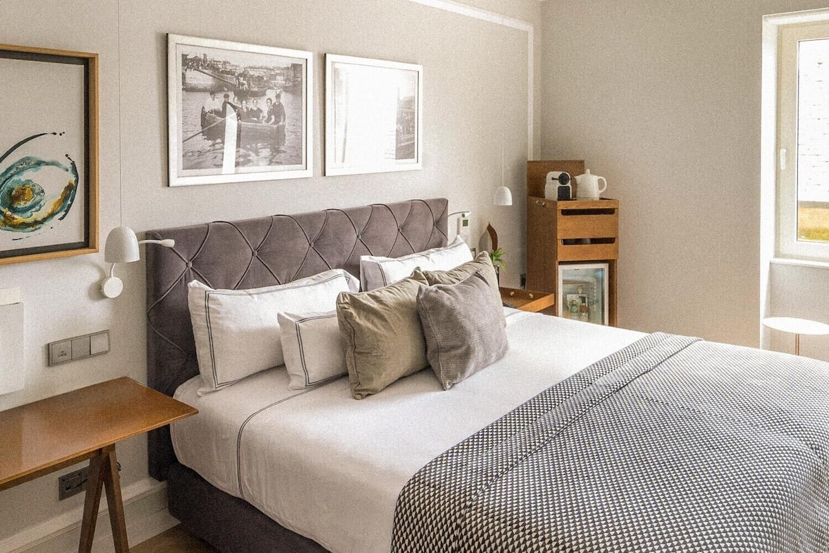 A bedroom with a bed, bedside tables, a nightstand, wall-mounted lamps, framed pictures, and a window.