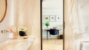Un baño con espejo, lavamanos y toallero. Se ve una pequeña mesa con un jarrón de flores y una silla al fondo.