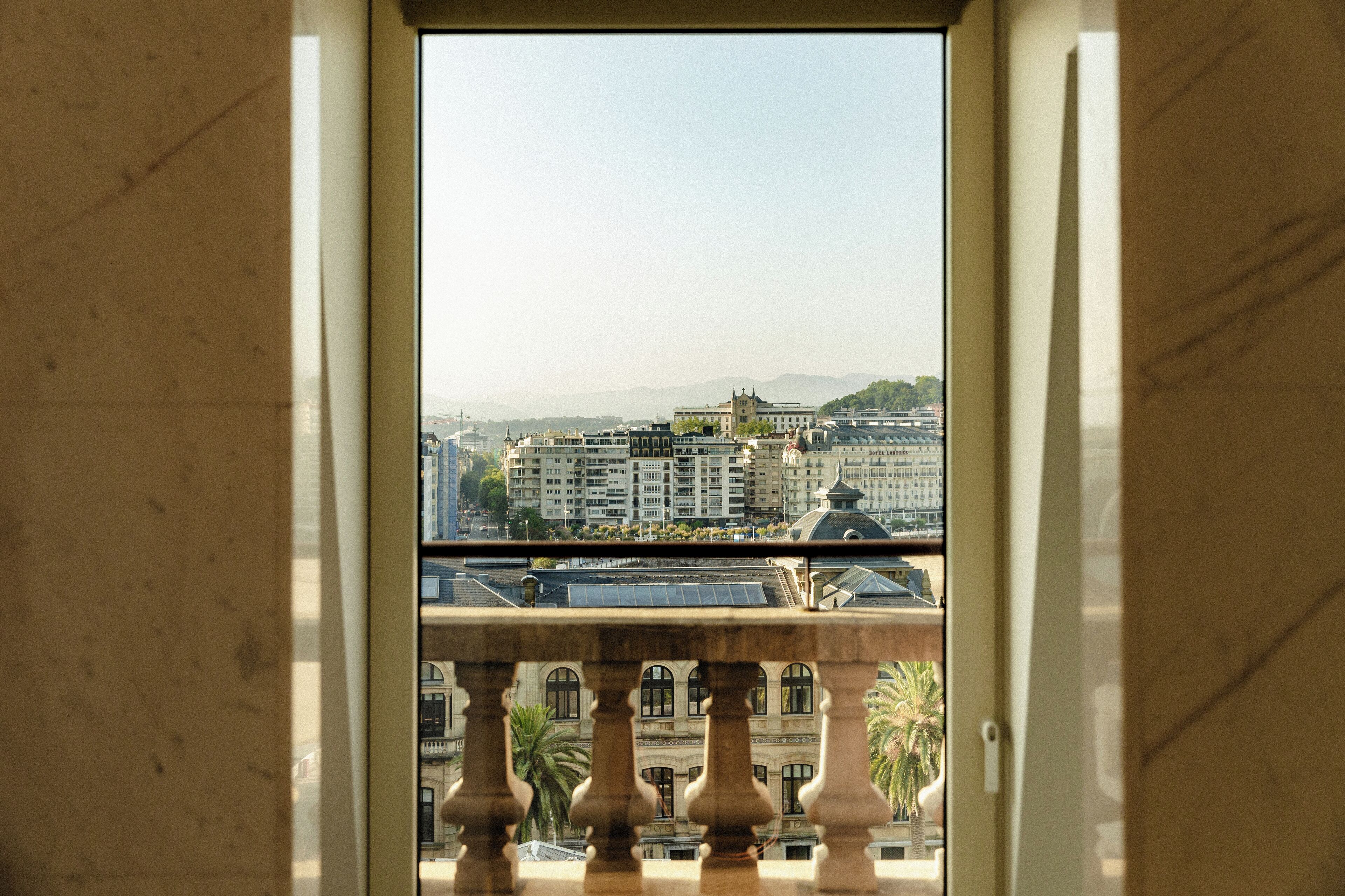 A view from a balcony with a marble balustrade, overlooking a cityscape with buildings and a distant mountain range.