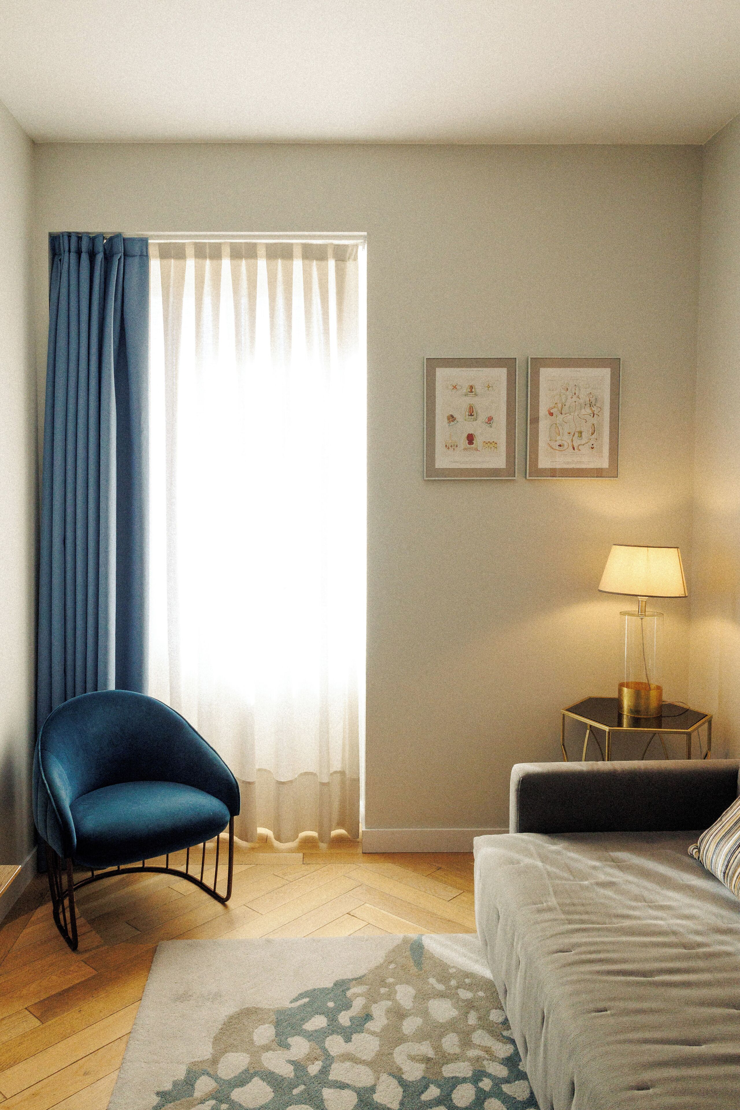 Un dormitorio moderno con un sillón azul, una cama, una mesita de noche con una lámpara y dos cuadros en la pared.