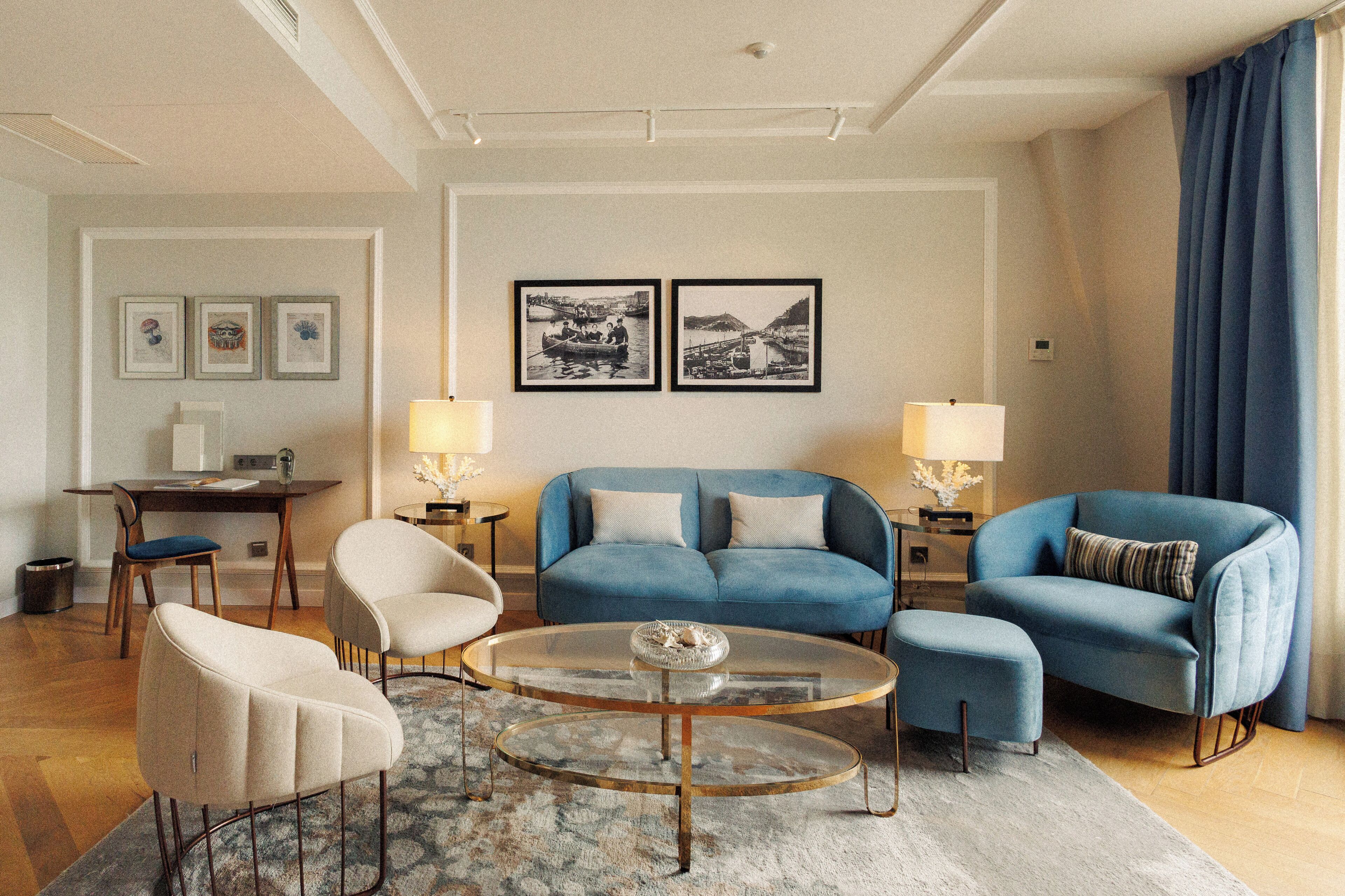 Una sala de estar moderna con un sofá azul, dos sillones, una mesa de centro de vidrio y un escritorio con una lámpara. Hay cuadros enmarcados en la pared y una ventana con cortinas.