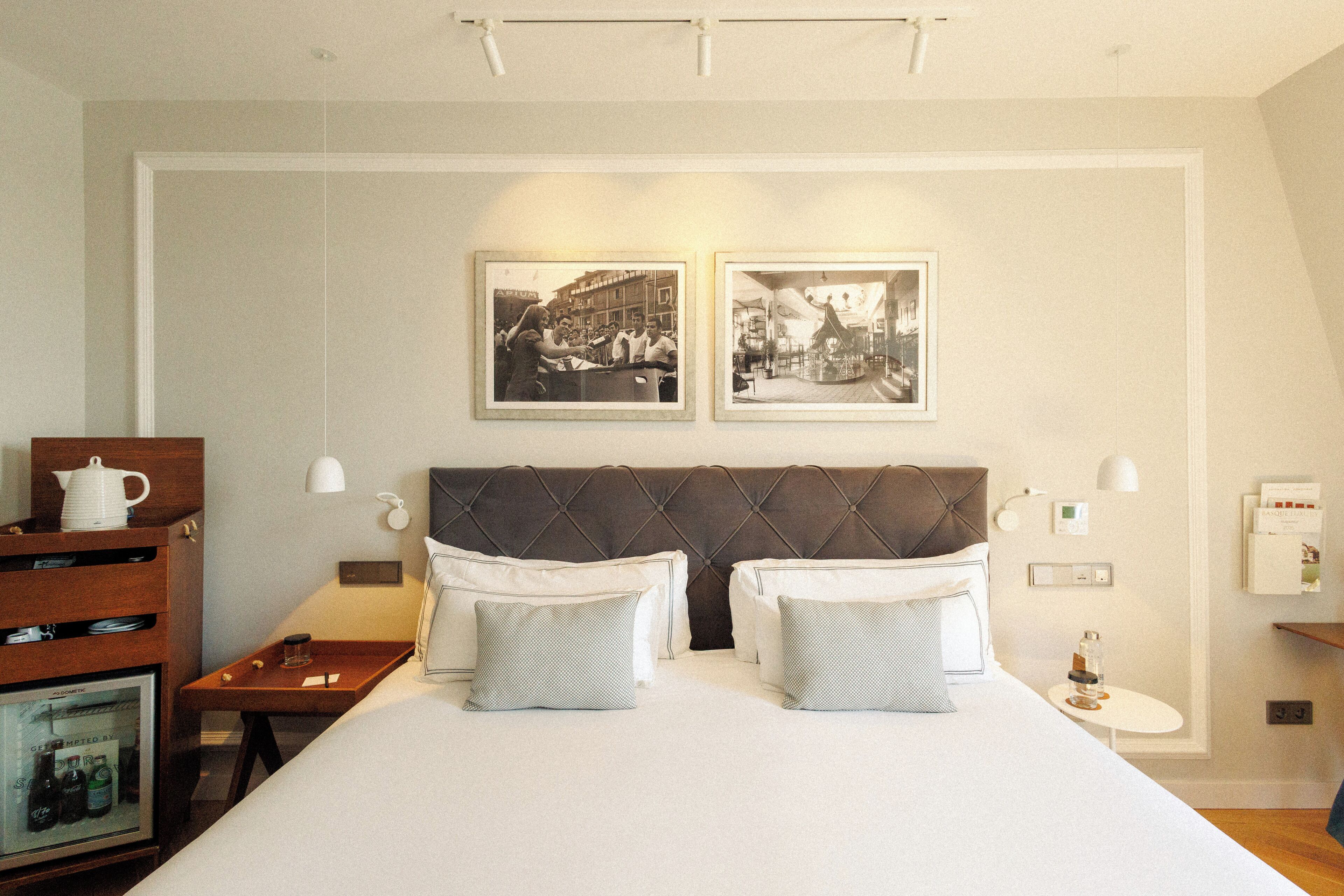 Quarto de hotel com uma cama grande, duas mesas de cabeceira, um aparador de madeira e duas fotografias emolduradas na parede.