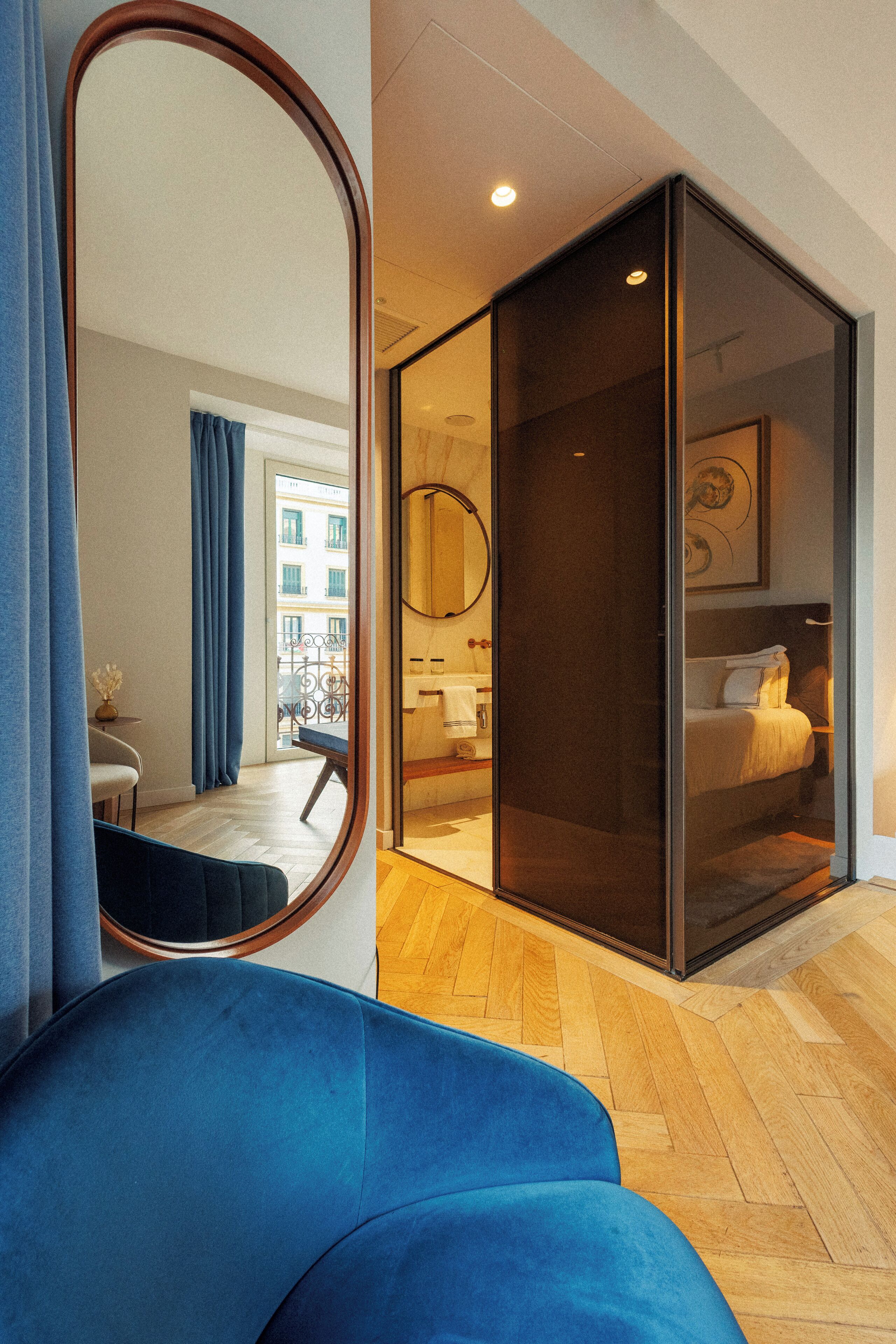 A modern hotel room with a large mirror, a blue sofa, a wooden herringbone floor, a sliding glass door leading to a balcony, and a bed with a white headboard.