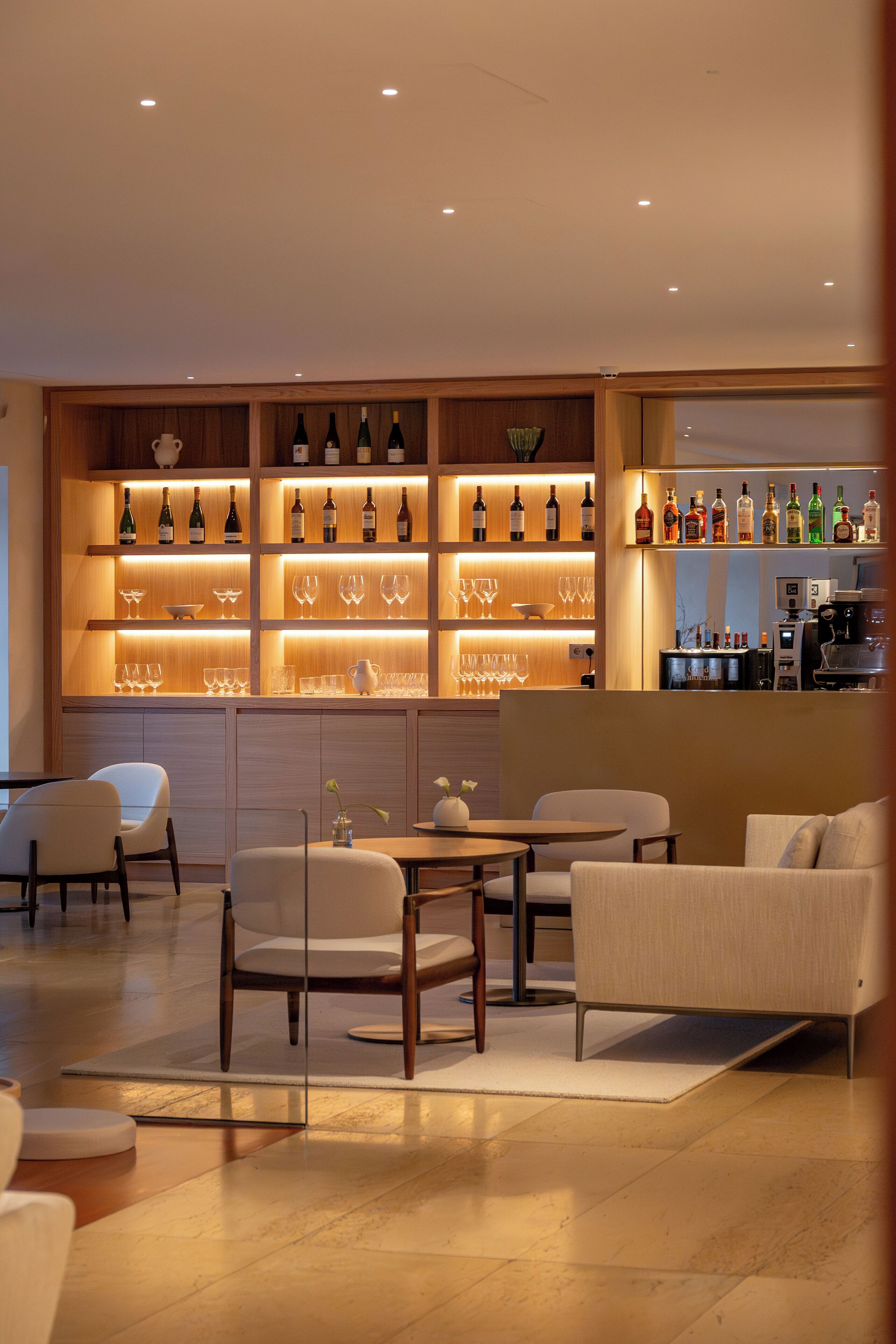 Un área de bar moderna con estantes llenos de botellas y vasos, una máquina de café y asientos sobre un piso de madera.