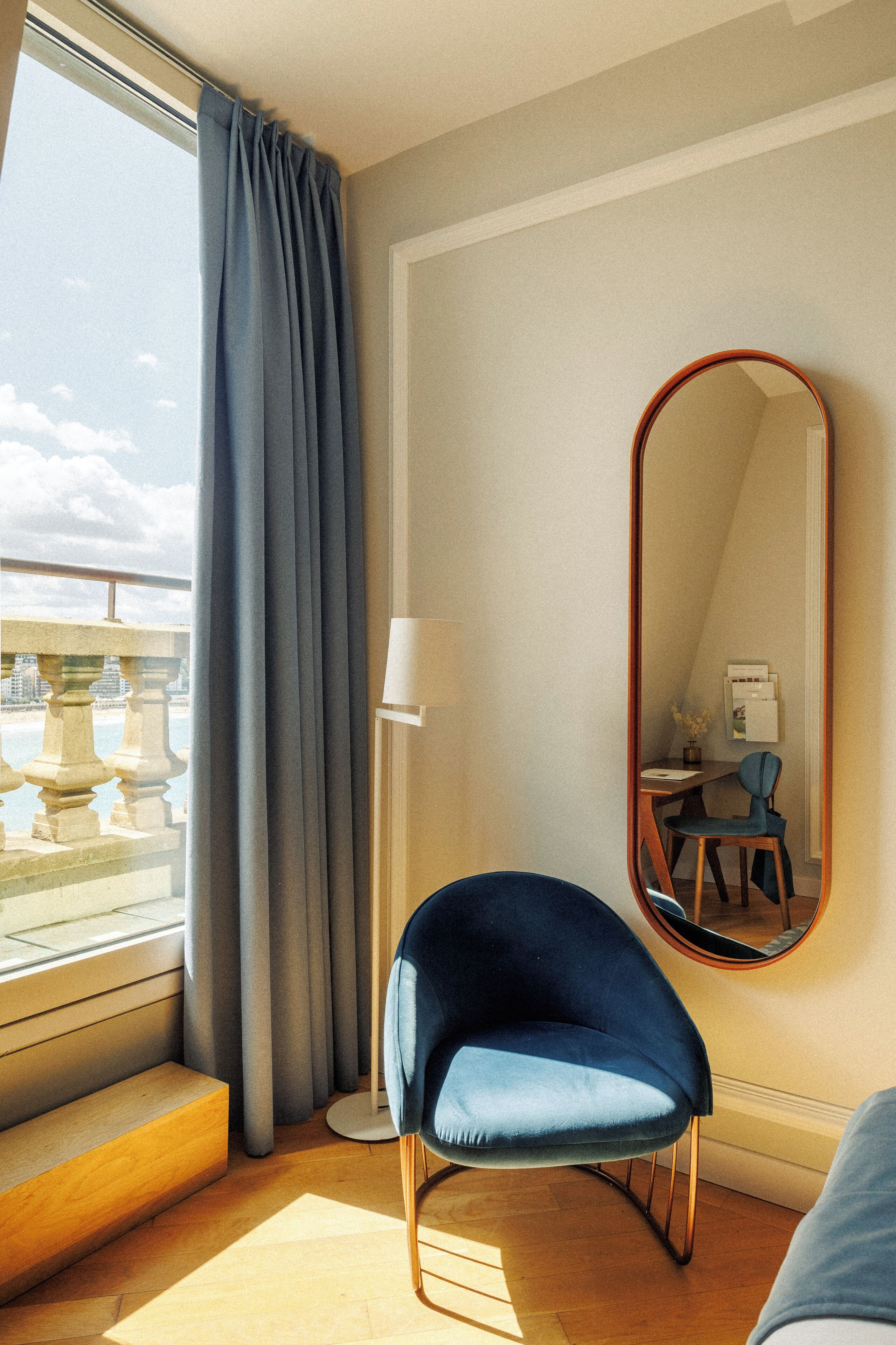 Una habitación con un ventanal grande, una silla azul, un espejo redondo y un escritorio con lámpara.