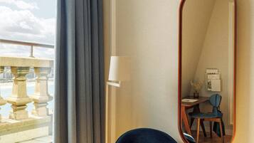 Una habitación con un ventanal grande, una silla azul, un espejo redondo y un escritorio con lámpara.