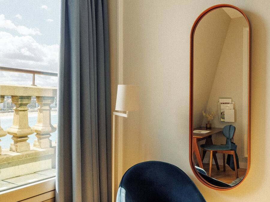 Una habitación con un ventanal grande, una silla azul, un espejo redondo y un escritorio con lámpara.