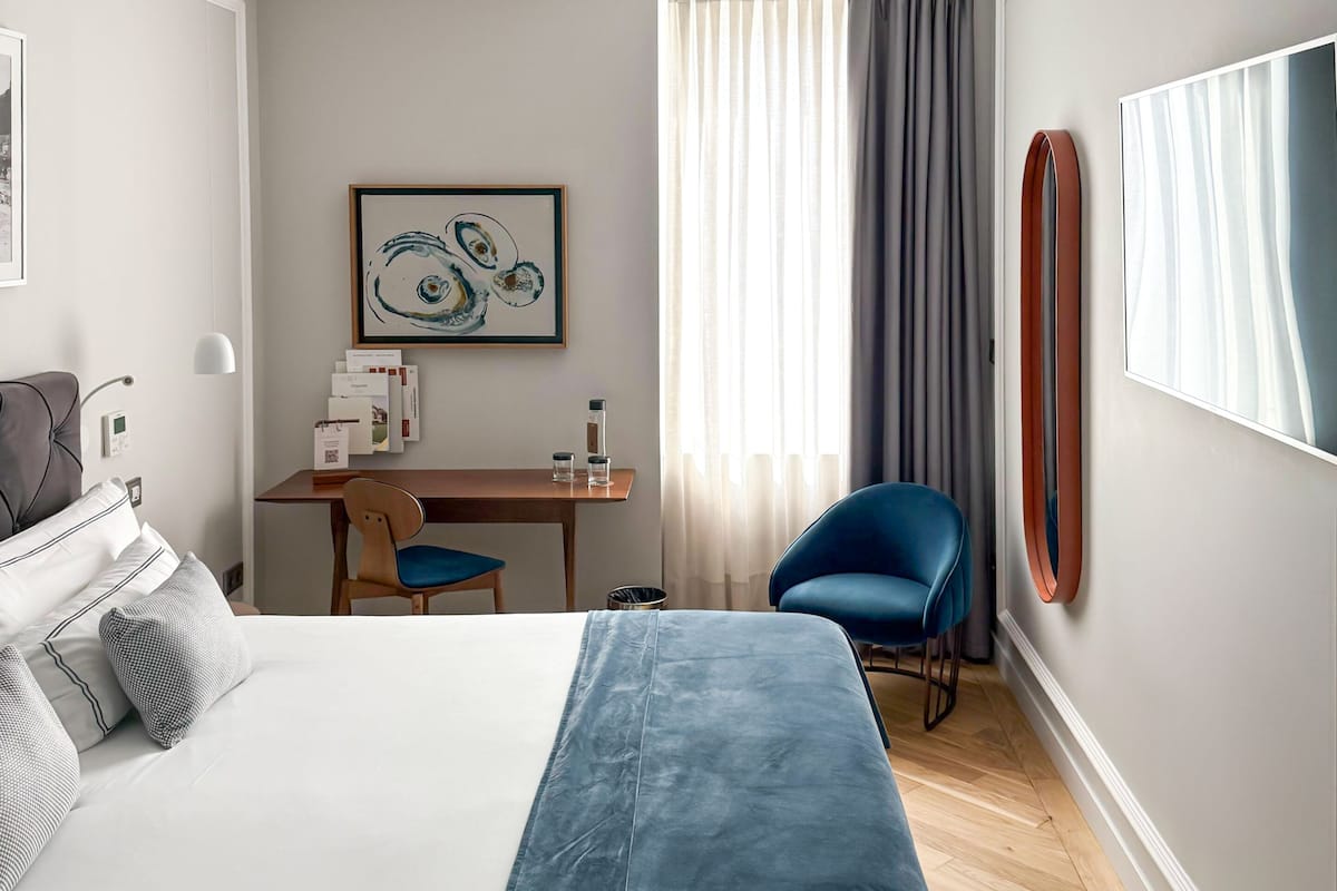 A hotel room with a bed, a desk, a chair, a large mirror, and a window with curtains.