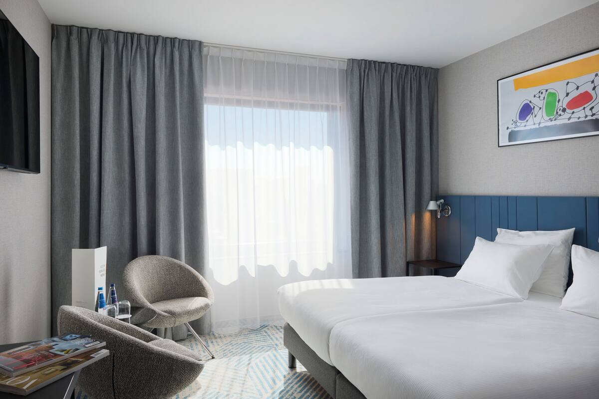 A hotel room with a large bed, a chair, a nightstand, and a window with curtains.