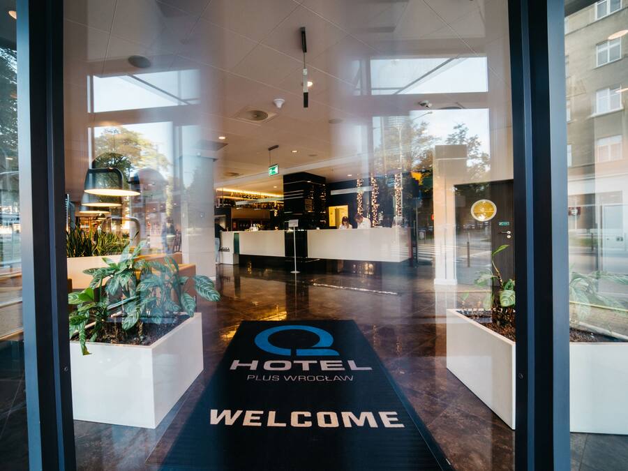 Zona de recepción de un hotel con un tapete de bienvenida, mostrador de recepción y plantas de interior.
