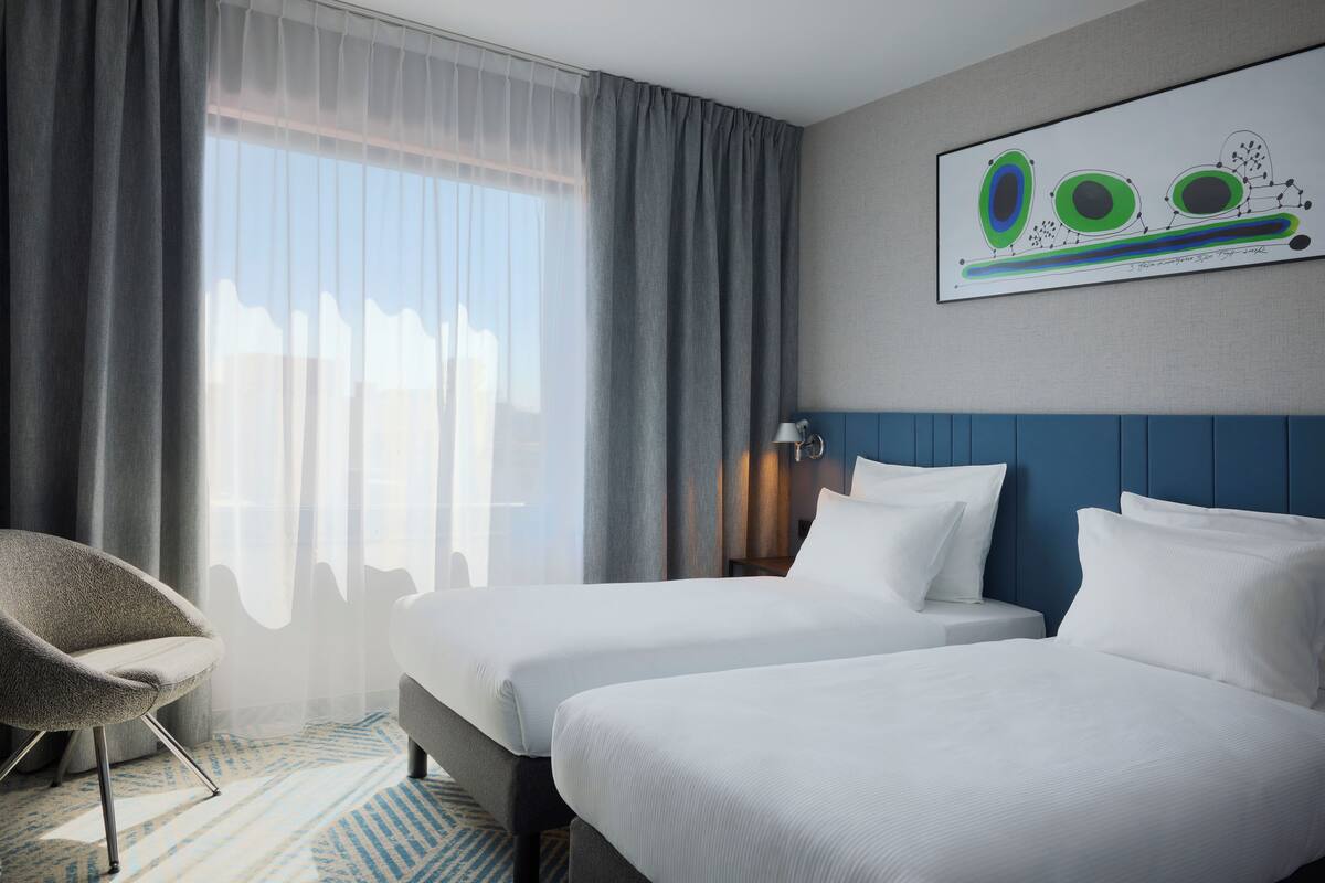 A hotel room with two beds, a chair, a painting, and a large window with curtains.