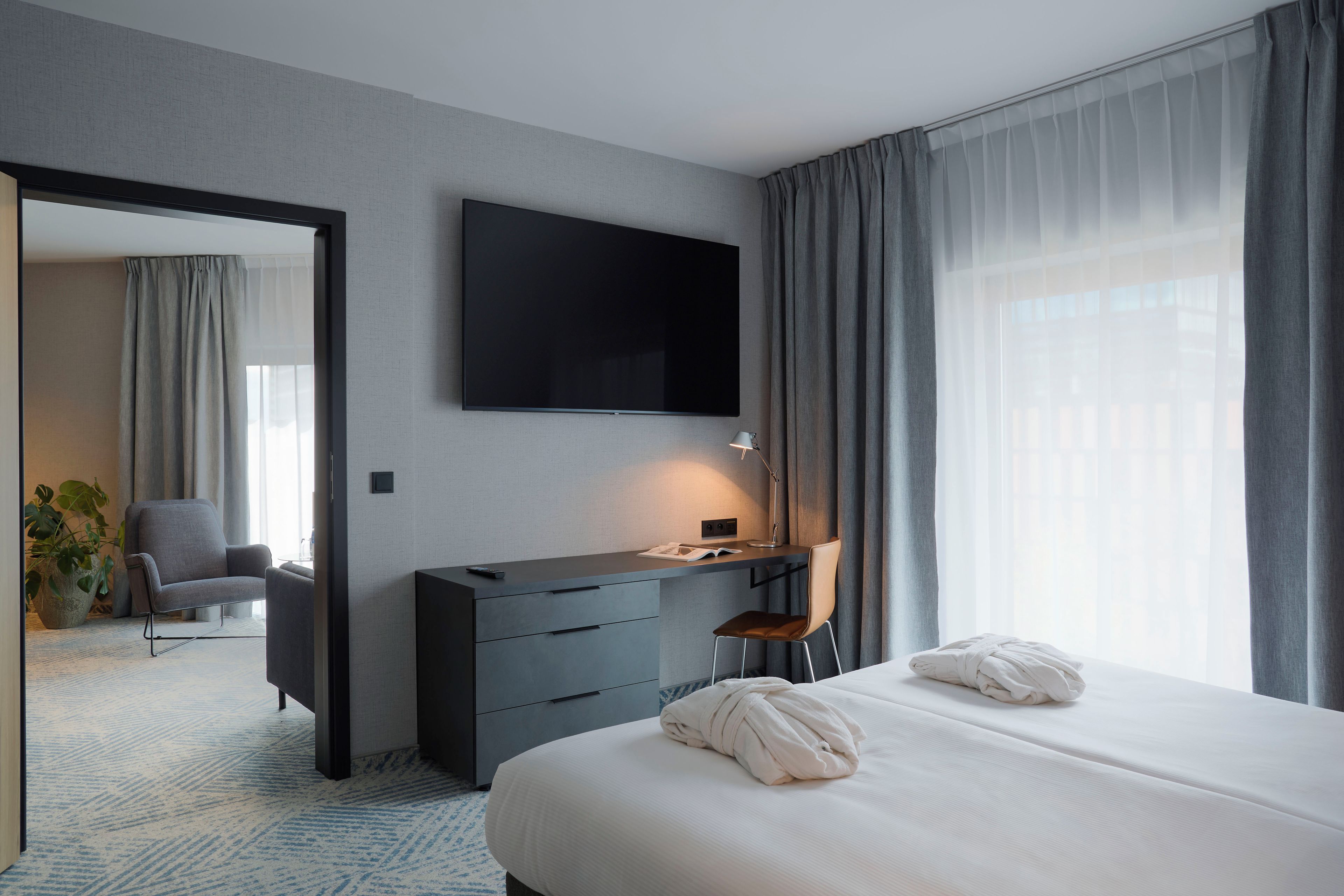 A hotel room with a bed, a desk with a chair, a television mounted on the wall, and a large window with curtains.