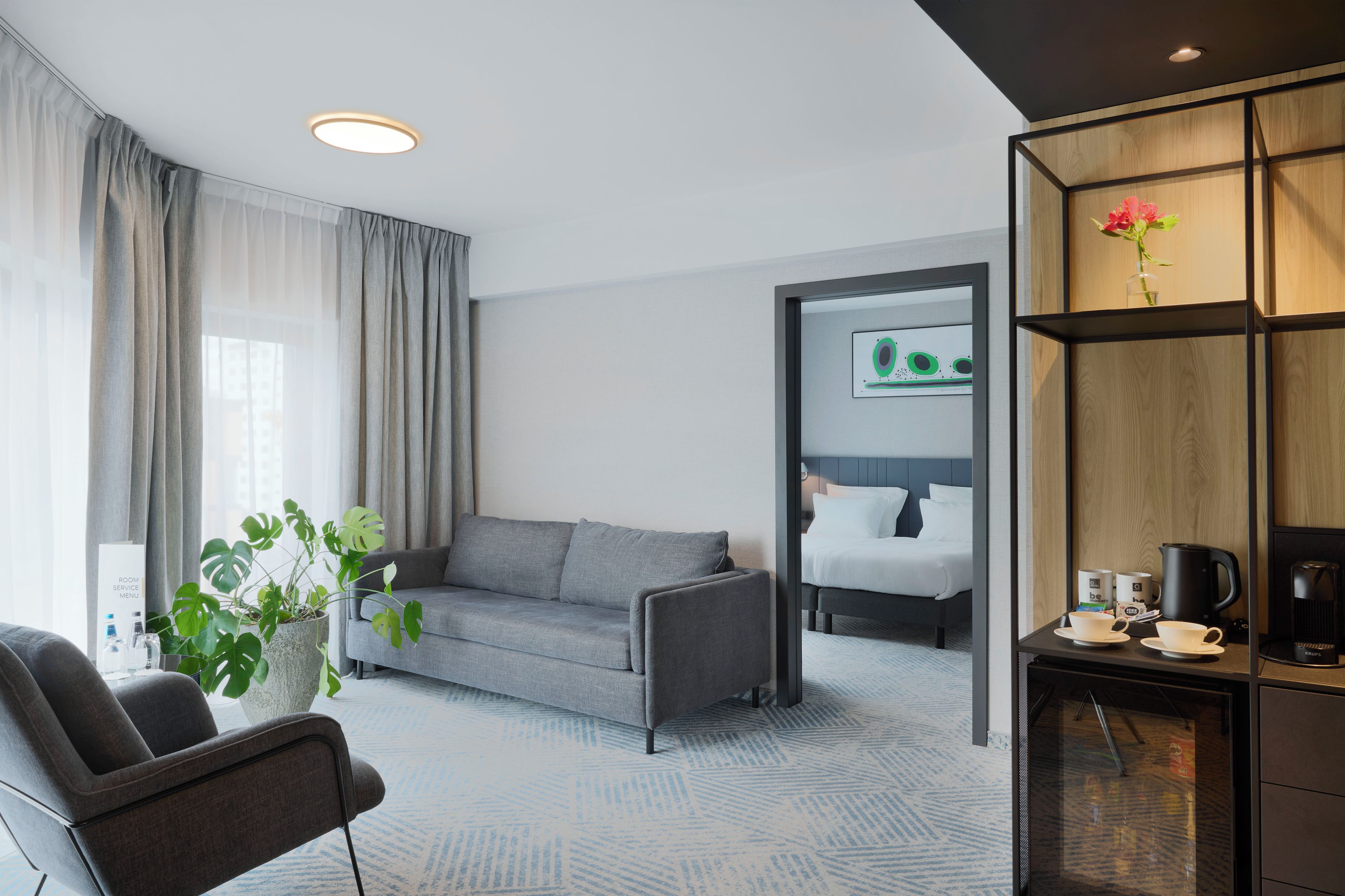 Een moderne hotelkamer met een grijze bank, een tafeltje met een koffiezetapparaat en een potplant.