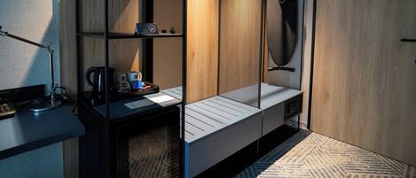 A modern hotel room with a wooden wardrobe, a desk with a kettle, and a hanging coat rack.
