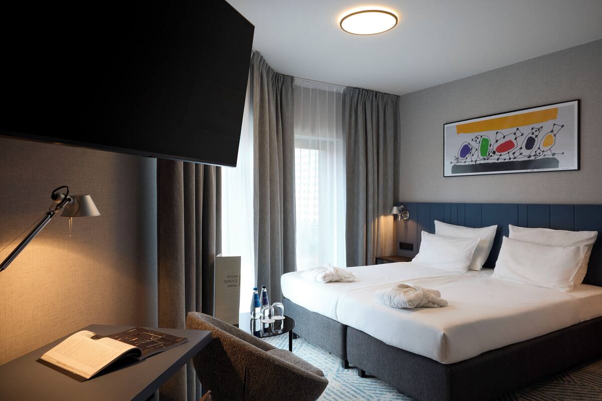 A hotel room with a bed, a desk with a lamp, a chair, a window with curtains, and a wall-mounted artwork.