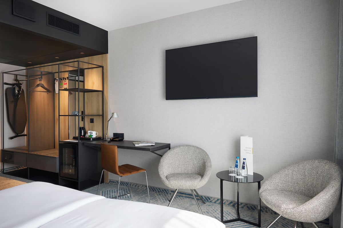 A modern hotel room with a bed, a desk, two chairs, a TV, and a mirror.