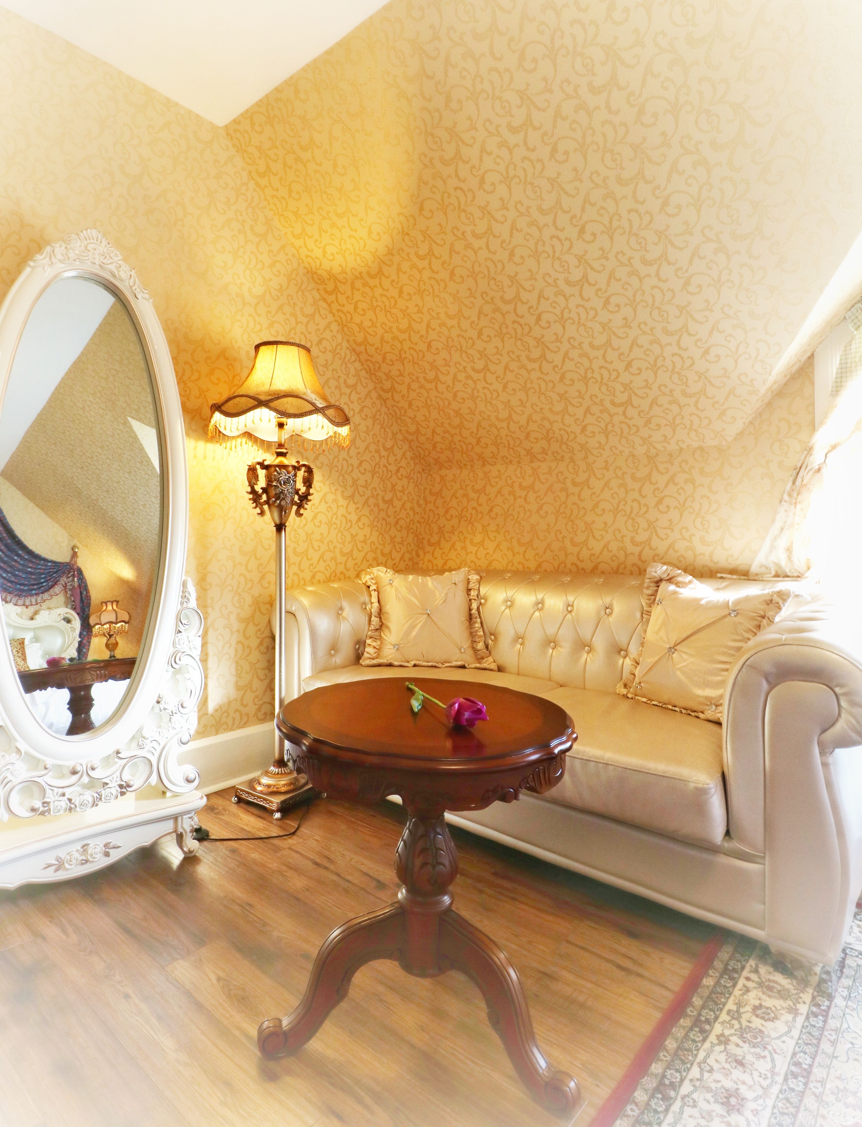 A room with a large ornate mirror, a round wooden table with a single purple rose, a cream-colored sofa, and a patterned wall.