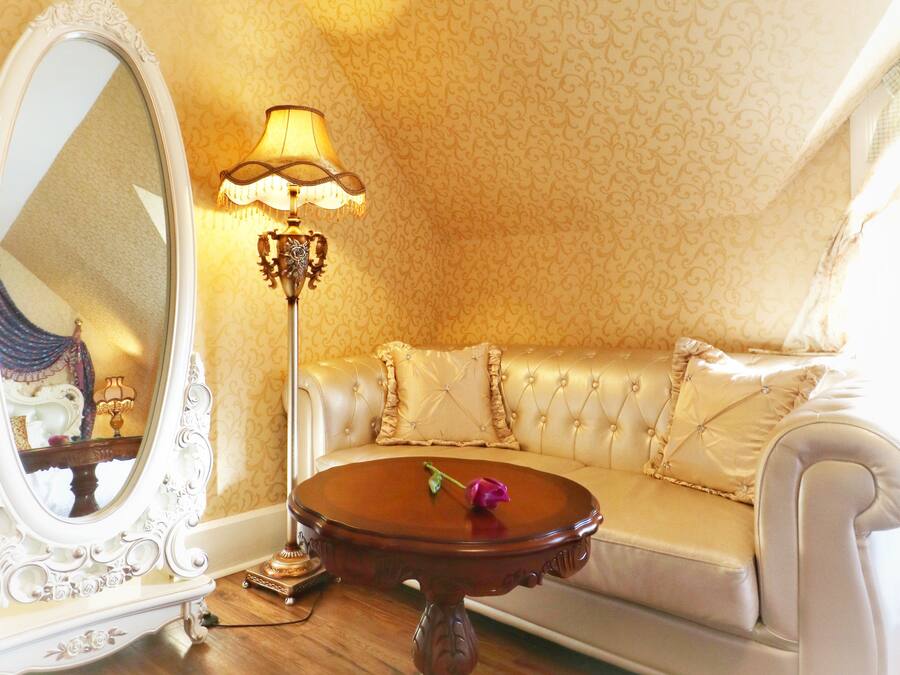 A room with a large ornate mirror, a round wooden table with a single purple rose, a cream-colored sofa, and a patterned wall.
