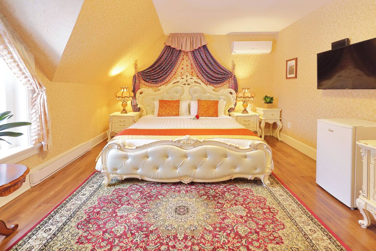 A bedroom with a large bed, ornate curtains, a television, and a small refrigerator.