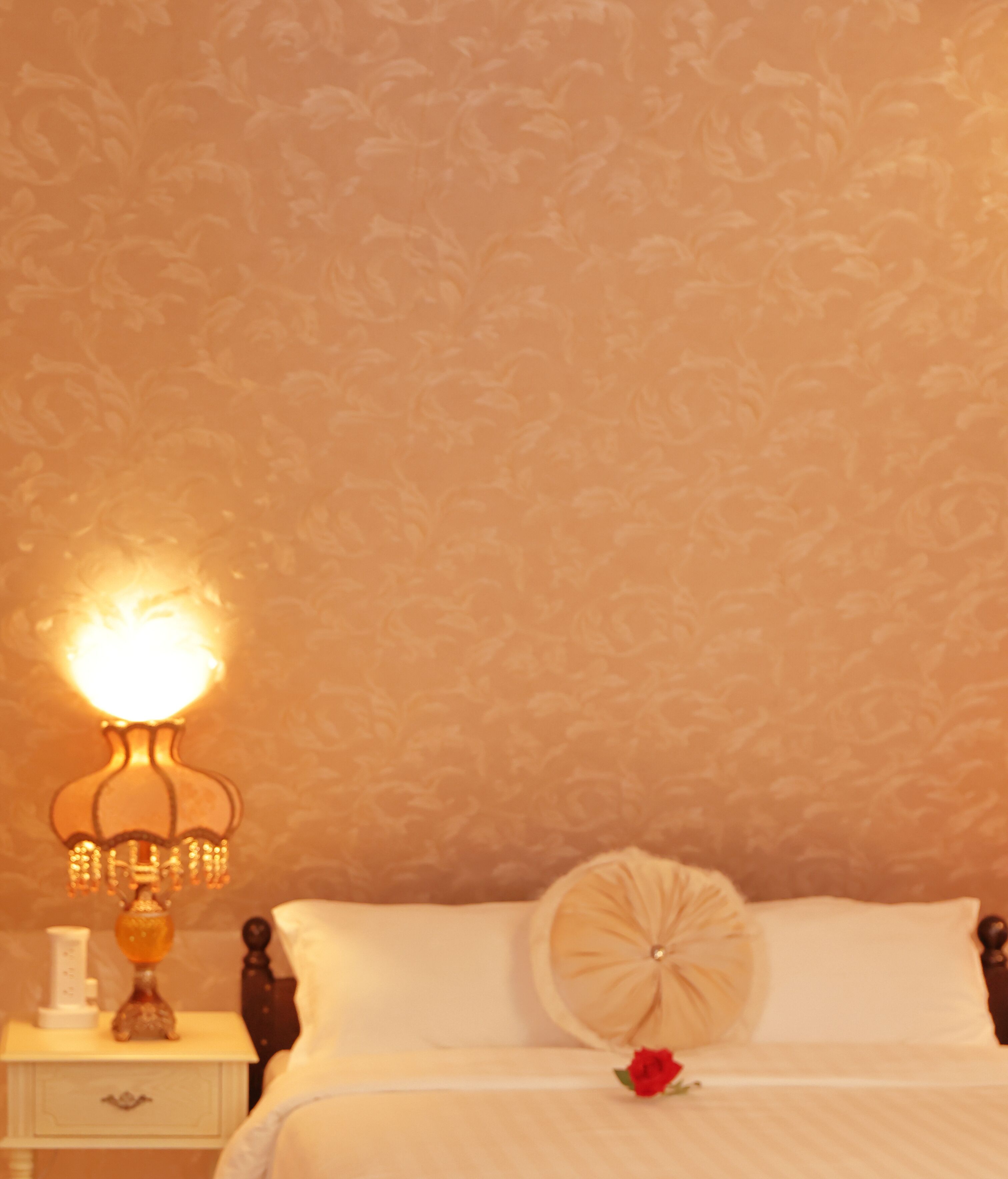 Een slaapkamer met behang met een patroon, een nachtkastje met een lamp, een bed met een kussen en een roos, en een nachtkastje met een fles.