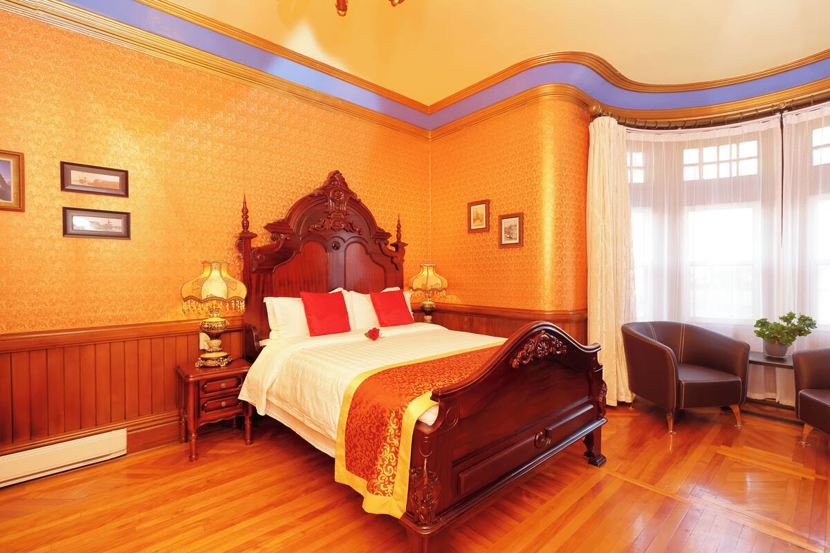 A room with a large wooden bed, two bedside tables with lamps, a chair, and a window with curtains.