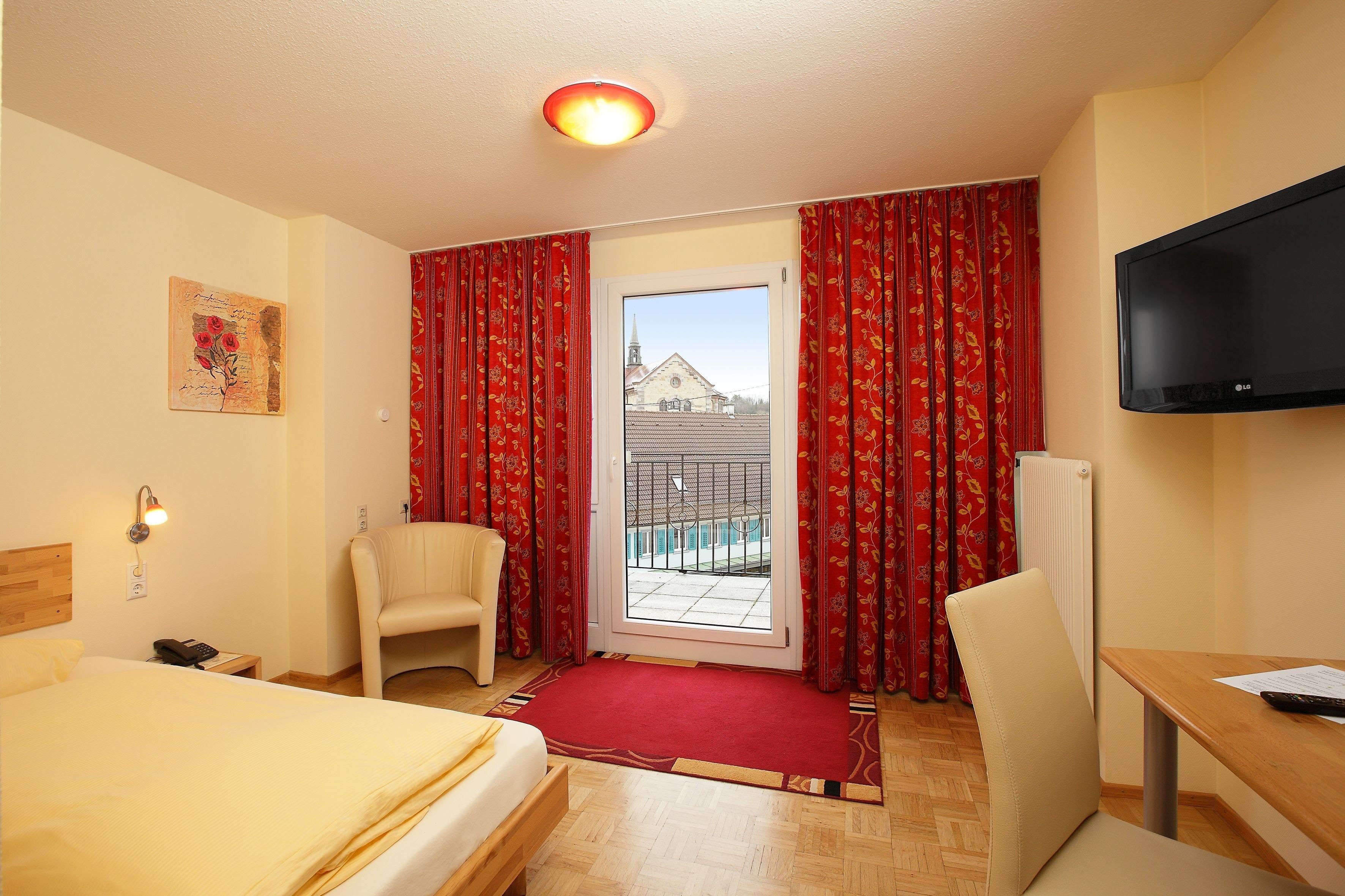 A hotel room with a bed, a chair, a table, a TV, and a view of a building through a door.