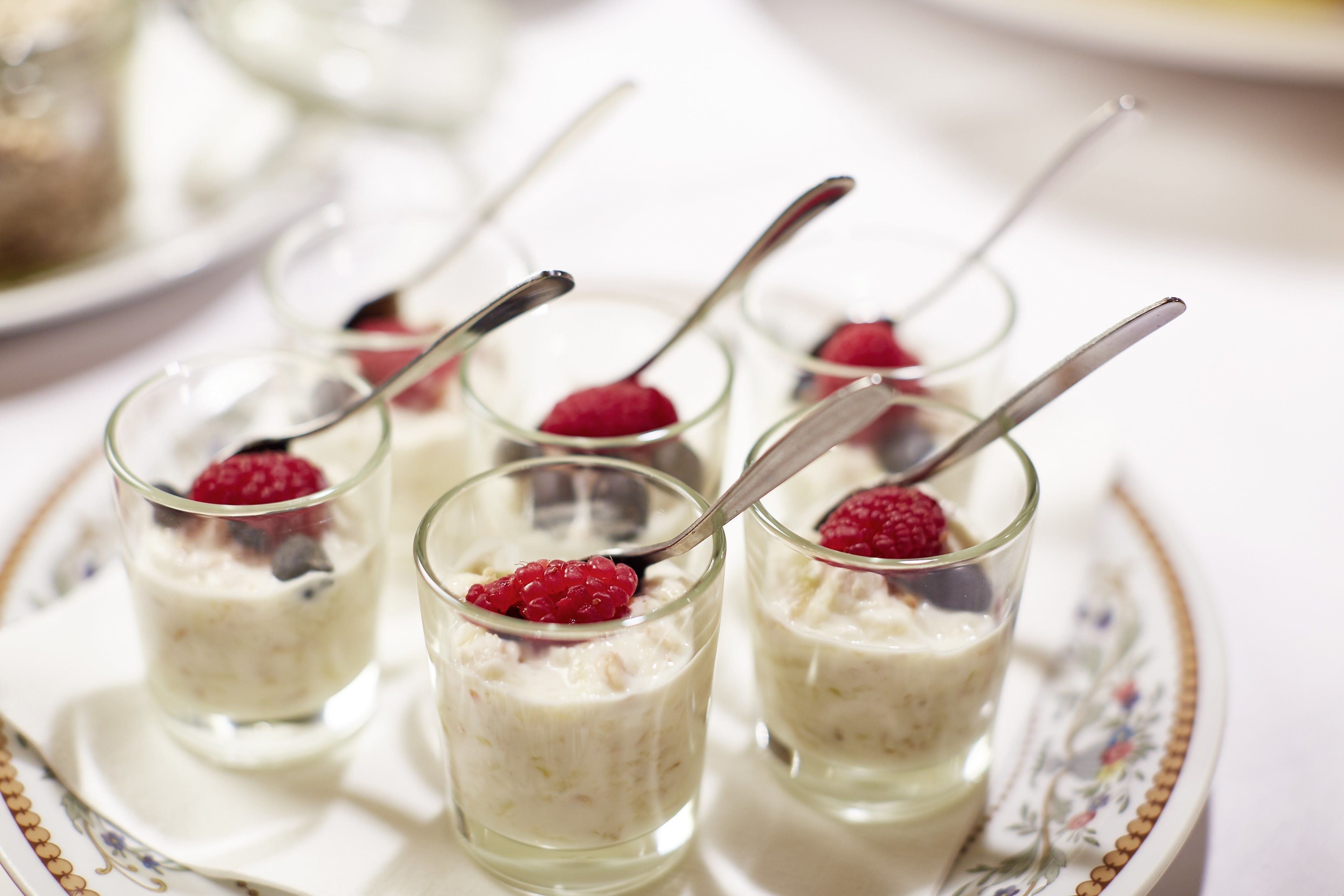 Vier Dessertgläser mit Löffel, jeweils mit einer Himbeere belegt, auf einer verzierten Platte.