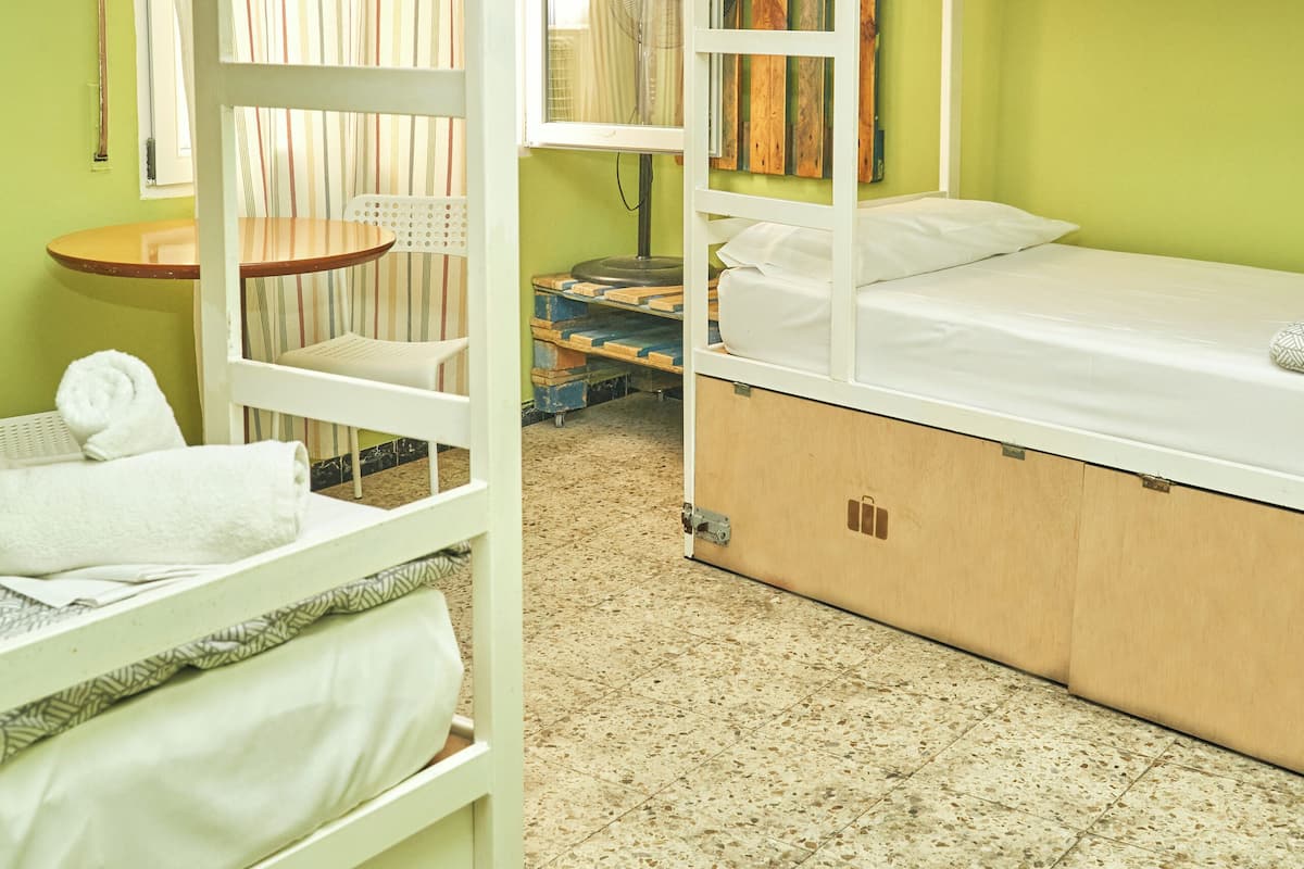 Een studentenkamer met stapelbedden, een stoel, een klein tafeltje en een ventilator.