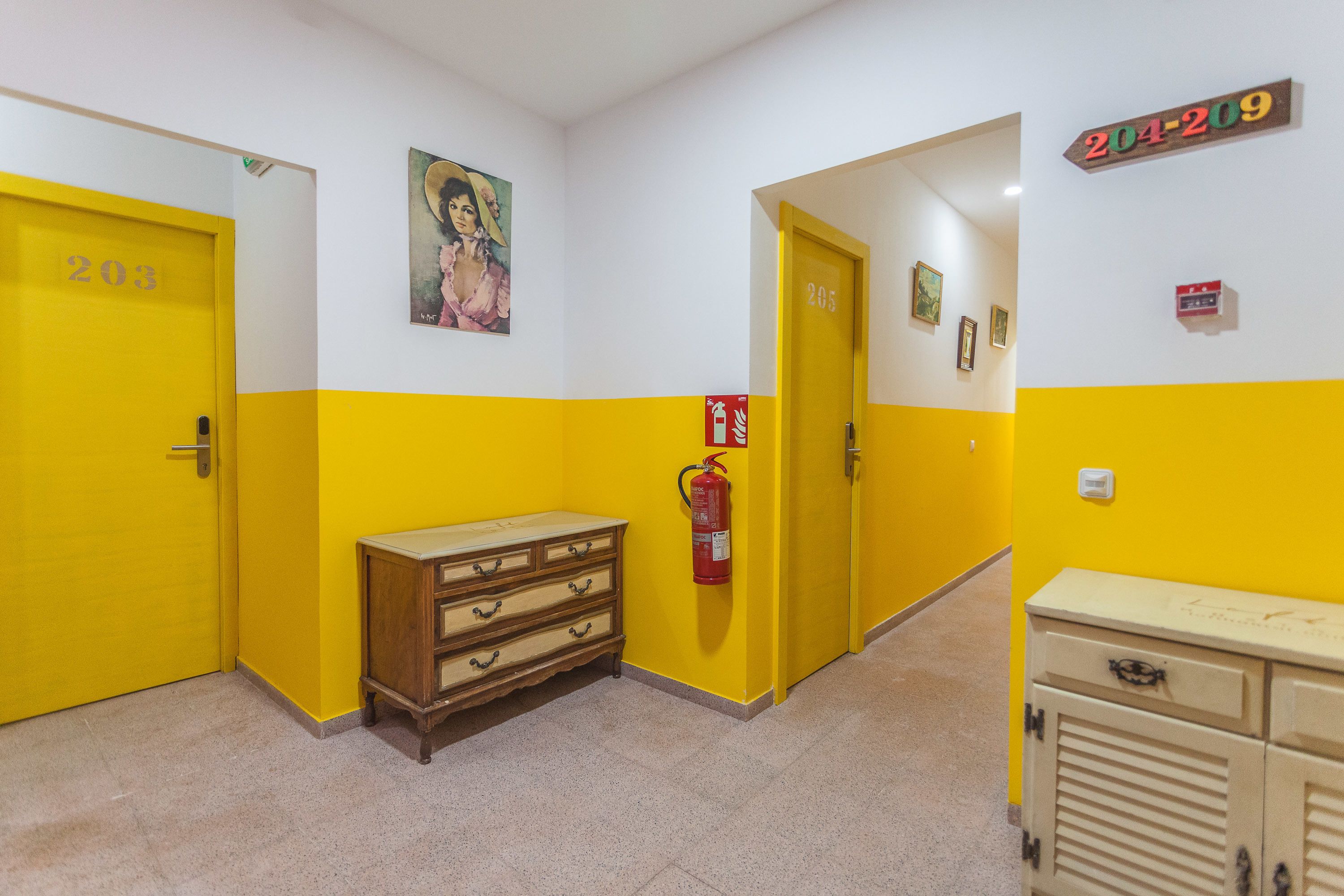 A hallway with yellow and white walls, two doors labeled 203 and 205, a wooden dresser, a fire extinguisher, and a cabinet.