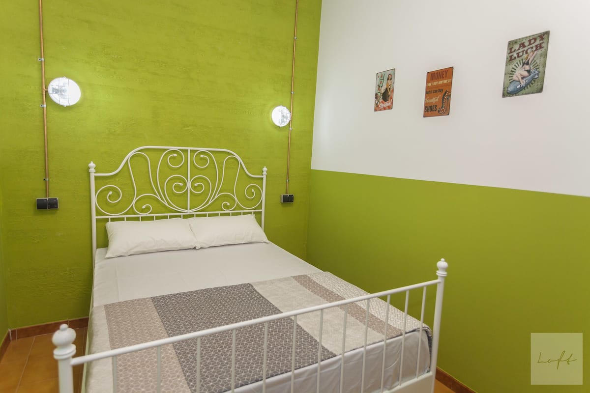 Een bed met een wit metalen frame en een geborduurd beddegoed, een groene wand en twee wandlampen.