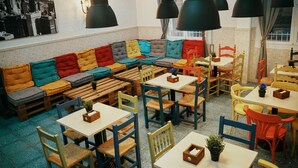 Een gezellig café met een kleurrijke bank, houten tafels en stoelen, en zwarte hanglampen.