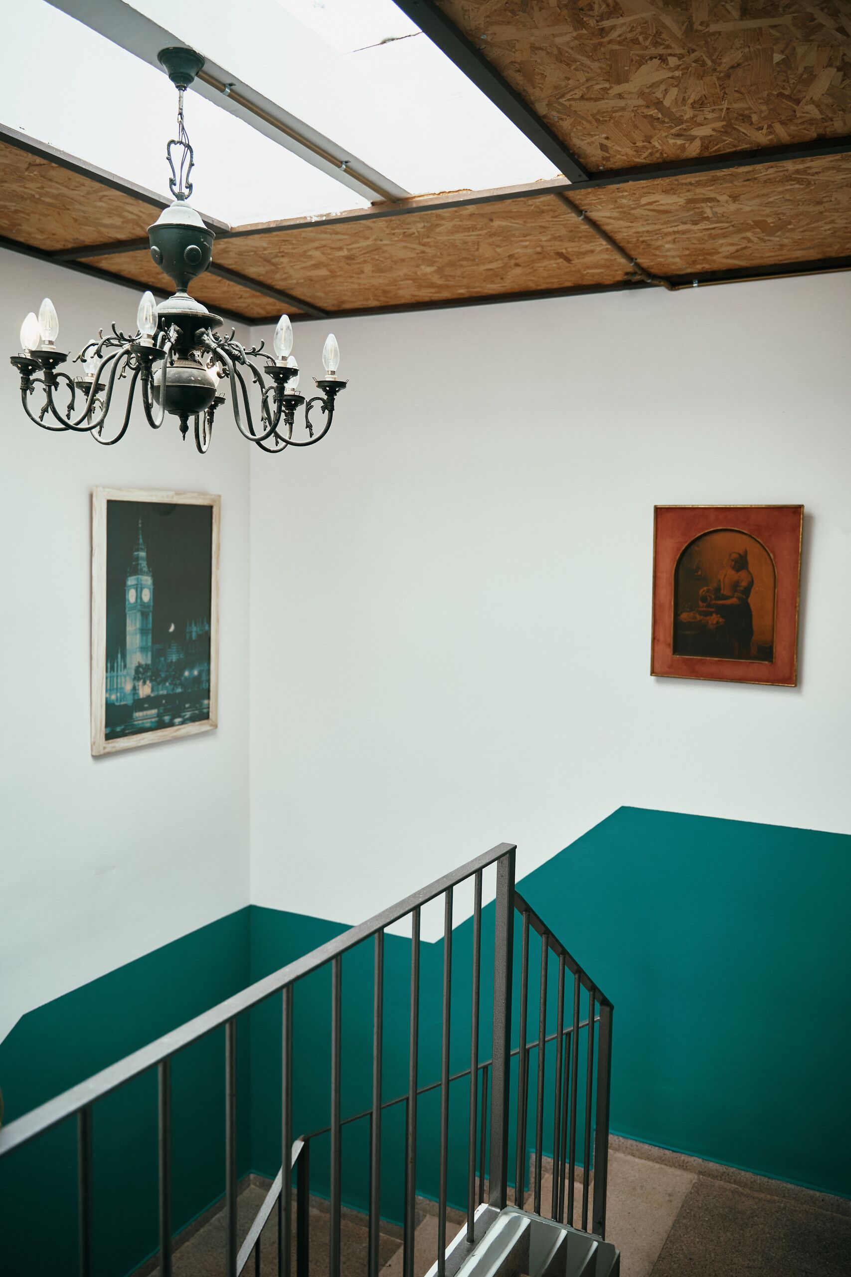 A staircase with a metal railing, a chandelier, and two framed pictures on the wall.