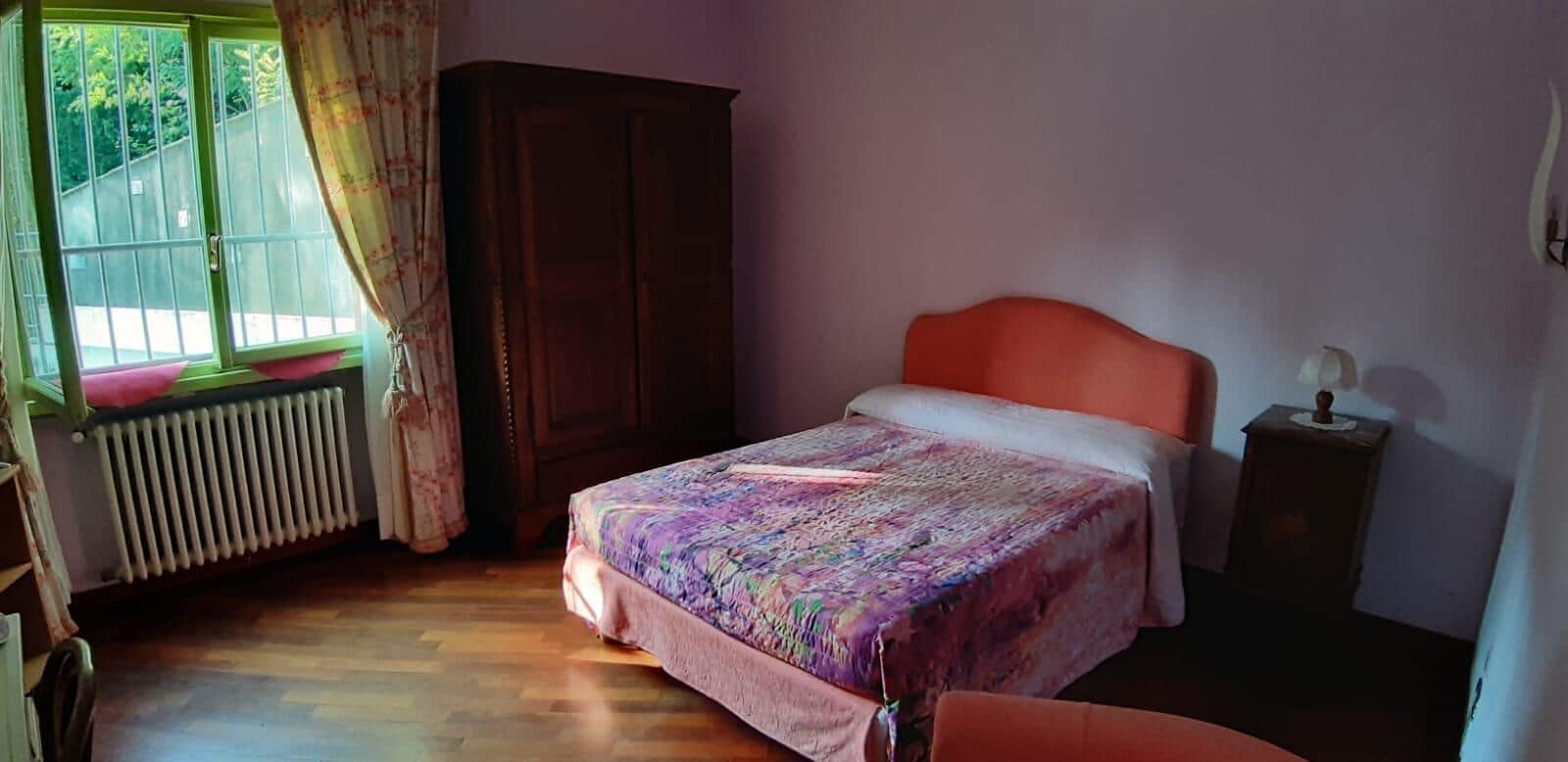 A bedroom with a bed, a radiator, a wardrobe, and a window with curtains.
