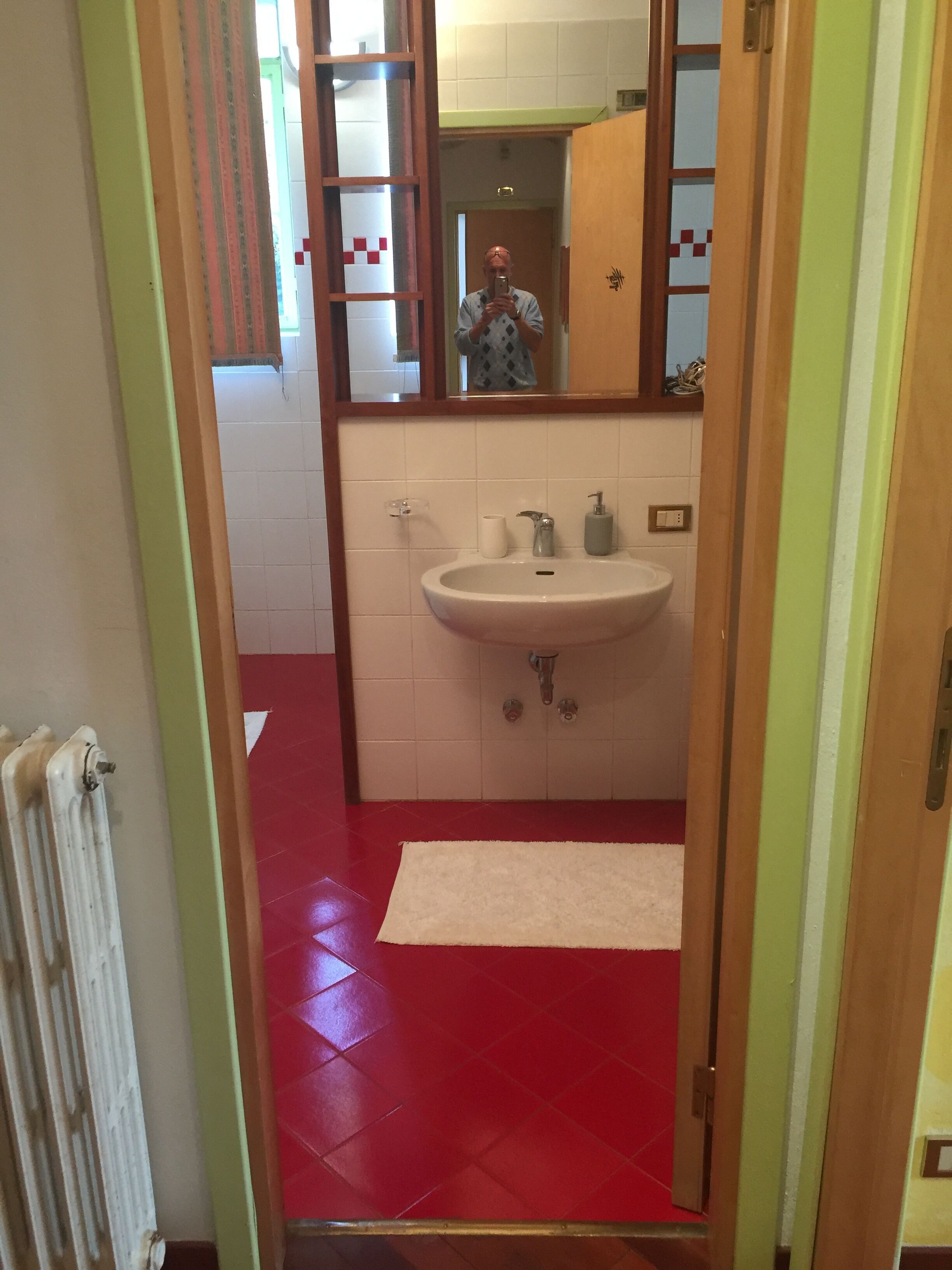 En smal gang med røde flisegulv, der fører til et badeværelse med en hvid vask, et spejl og en radiator.