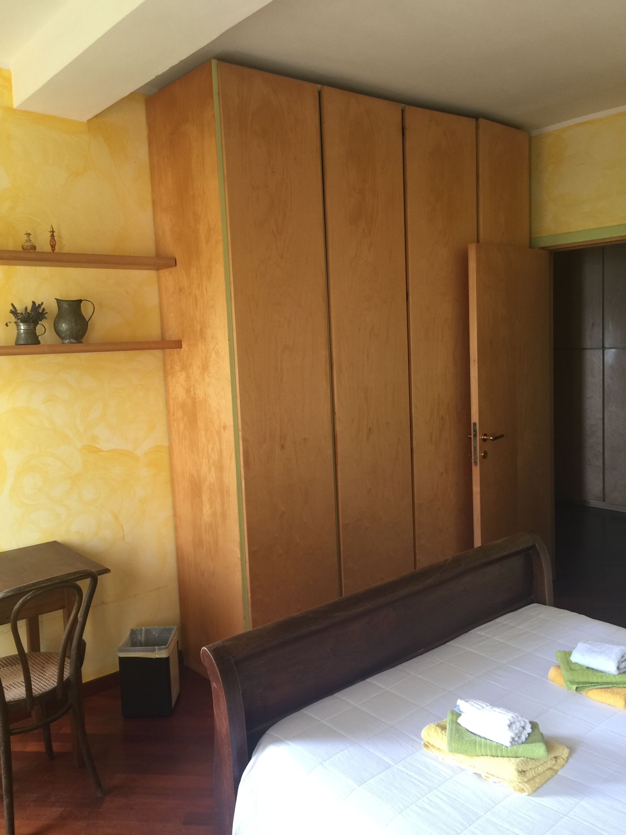A bedroom with a wooden wardrobe, a bed with towels, a small table, and a chair.