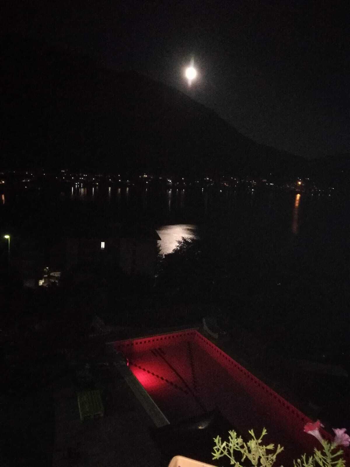 Een nachtzicht van een stadsbeeld met een grote maan op de hemel, een waterpartij die stadslichten weerspiegelt, en een rood verlichte constructie op de voorgrond.