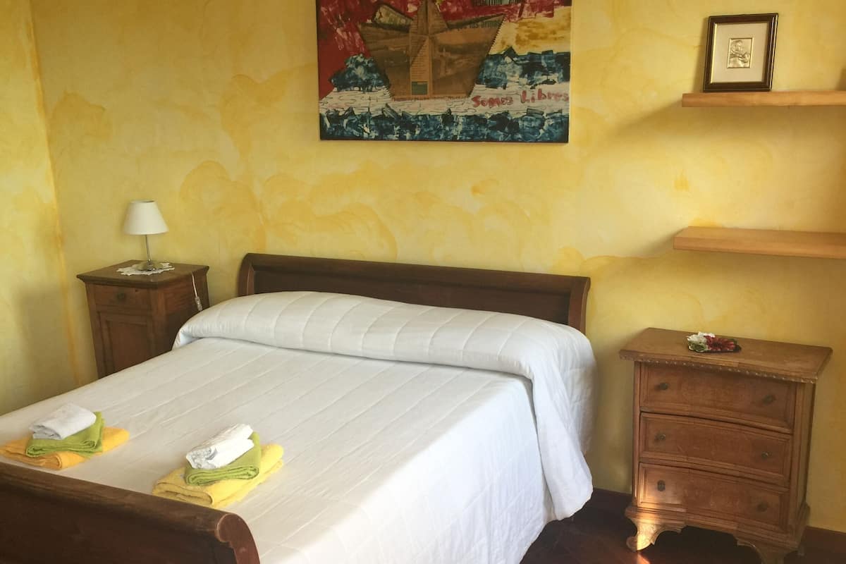 A neatly made bed with a white bedspread, a wooden nightstand with a lamp, a small wooden dresser, and a wall with a colorful painting.