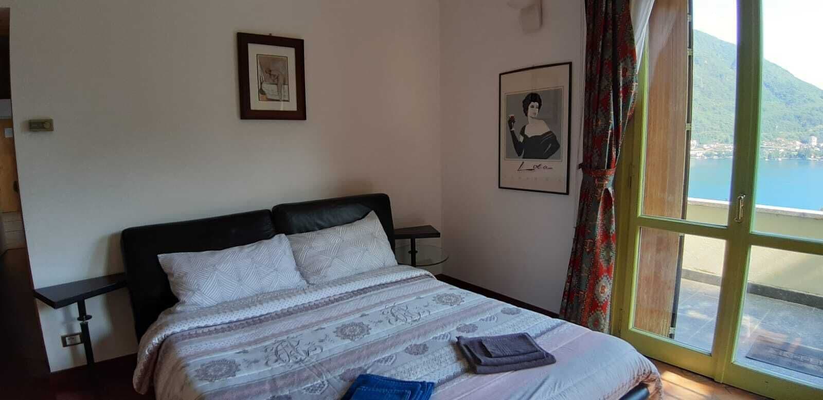 A bedroom with a bed, a nightstand, a window with a view of a body of water and hills, and two framed pictures on the wall.