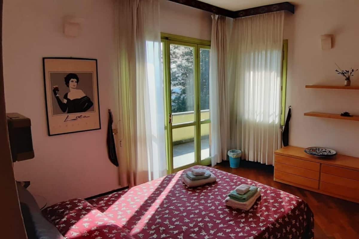 A bedroom with a bed, a dresser, a window with curtains, a framed picture on the wall, and a small shelf with decorative items.