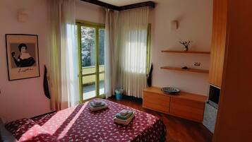 Een slaapkamer met een bed, een dressoir, een raam met gordijnen, een lijst met een afbeelding aan de muur en een klein plankje met decoratieve objecten.